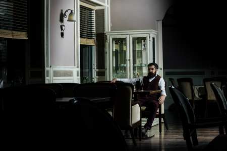 Man sitting at table served with bottle of wine and glasses in restaurant. Brutal hipster with beard in brown vest and trousers. Alcohol and appetizer. Unhealthy lifestyle and bad habitsの写真素材