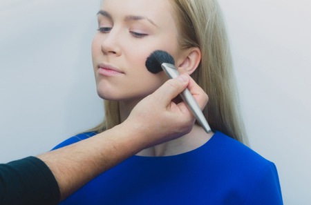 Girl getting powder on face with makeup brush in male hand. Adorable woman with young healthy skin and blond long hair on grey background. Visage, cosmetics, make up and skincare. Beauty salonの写真素材