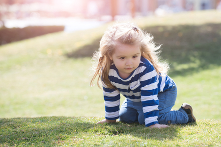 Boy little child with adorable thoughtful face with long blond hair in blue and white striped shirt and jeans crawling on his knee on meadow with green grass in summer dayの写真素材