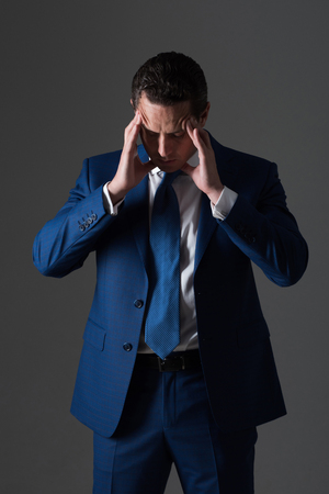 Tired boss, handsome man or businessman feeling headache with stylish hair, haircut in fashionable blue formal suit, white shirt and tie on grey background. Business. Stress and problemsの写真素材