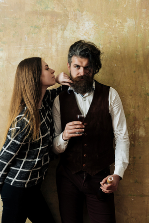Woman with hipster holding bottle and glass of wine. Girl with long hair and man with beard and messy haircut on beige wall. Couple in love. Alcohol and convive. Unhealthy lifestyle. Bad habitsの写真素材