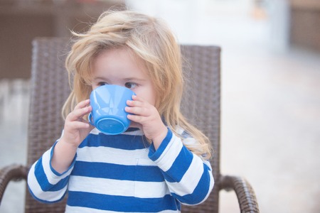 Boy drinking milk. Small child with blonde hair in striped blue and white shirt drinking milk from cup sitting in brown armchairの写真素材