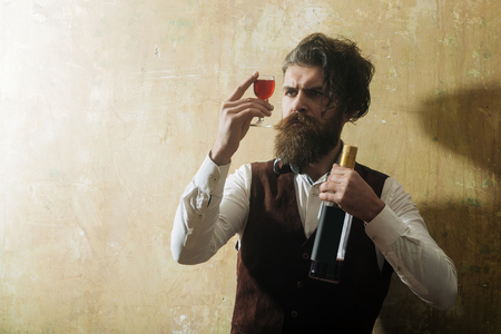 Man with bottle looking at red wine in glass. Brutal hipster with long beard, moustache and messy hair on beige wall. Alcohol abuse and alcoholism. Unhealthy lifestyle. Bad habits, copy spaceの写真素材