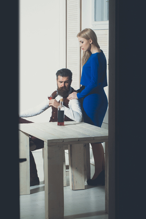 Girl standing at man drinking wine. Sexy woman with blond hair in blue dress and bearded hipster with bottle on wooden table. Couple in love. Alcohol and convive. Unhealthy lifestyle. Bad habitsの写真素材