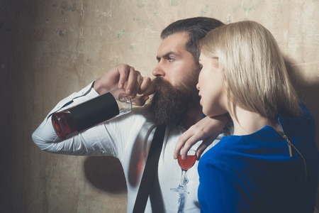 Couple in love with alcoholic drinks. Girl with glass of liqueur and man drinking wine from bottle. Woman with blond hair and bearded hipster on beige wall. Alcohol and convive. Unhealthy lifestyleの写真素材