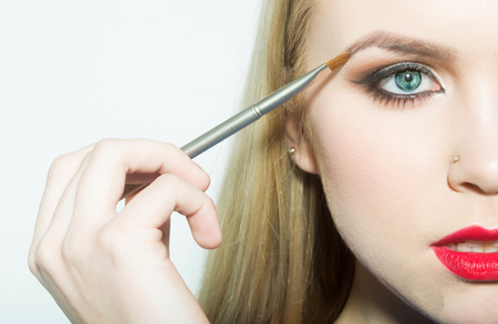 Girl half face drawing eyebrow with makeup brush. Woman with blue eyes, red lips makeup and blond long hair isolated on white. Beauty salon. Visage and make up. Cosmetics and skincareの写真素材
