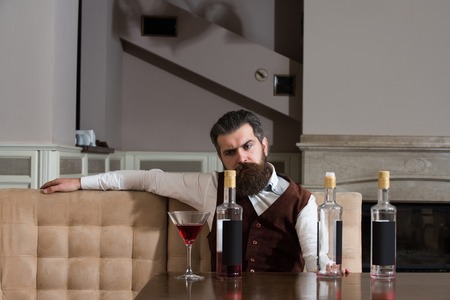 Man drinking red wine alone in restaurant. Bearded hipster with beard sitting at table with three bottles and glass. Alcohol abuse. Addictive and convive. Unhealthy lifestyle and bad habitsの写真素材