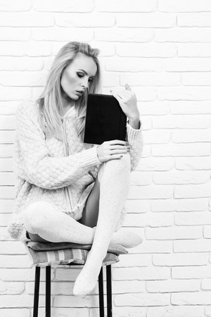 girl or beautiful cute woman fashion model with blond hair in sexy knitted sweater reads book sitting on chair on brick wall, black and whiteの写真素材