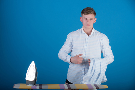 Macho wearing striped shirt. Housework and fashion concept. Man with laundry for ironing. Model using iron. Ironing board on blue background.の写真素材
