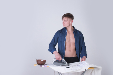 Macho in blue shirt with sexy torso ironing clothes with iron on grey background. Bowl with food for healthy dieting. Housework concept, copy spaceの写真素材