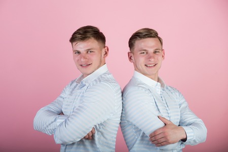 Two brothers smiling on pink background. Happy men with hands folded. Models standing together. Twins wearing blue shirts. Family, brotherhood and friendship concept.の写真素材