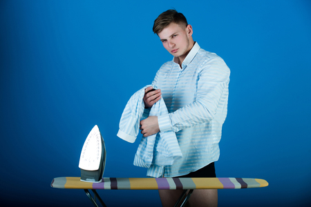 Man with laundry for ironing. Model using iron. Ironing board on blue background. Macho wearing striped shirt. Housework and fashion concept.の写真素材