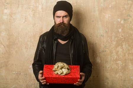 Holiday celebration and shopping. Hipster with present box in hat. Christmas and birthday. Guy with serious face and gift pack. Man with long beard.の写真素材