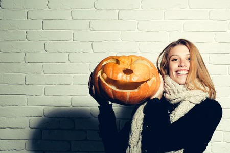 Halloween woman with happy face. Girl with orange scary pumpkin. Holiday and celebration. Woman in coat and scarf at brick wall. Party and traditional food, copy spaceの写真素材