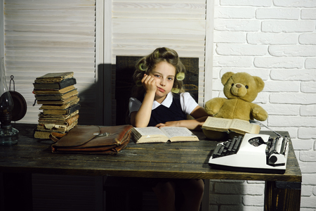 Small girl with curler in hair read book. Child with briefcase and typewriter on table. Education and childhood. Kid choose career of journalist or writer. Little baby secretary in cabinet or library.の写真素材