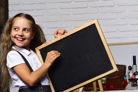 Schoolgirl with happy smile holds small blackboard. Girl in her classroom or laboratory with bag and stationery. Kid and school supplies on white brick background. Back to school and childhood conceptの写真素材
