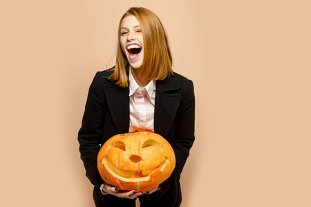 Halloween woman with happy face. Woman in coat and scarf at beige wall. Party and traditional food. Holiday and celebration. Girl with orange scary pumpkin.の写真素材