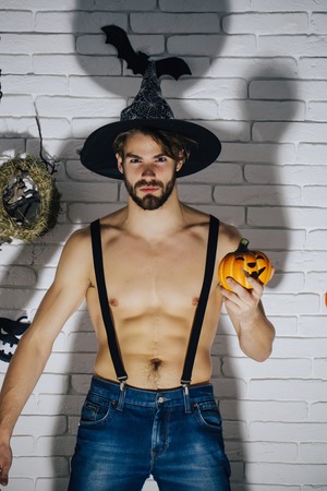 Halloween man in witch hat with pumpkin. Macho with muscular torso and dark shadow. Holiday traditional symbols on white brick wall. Jack o lantern. Mystery and horror concept.の写真素材