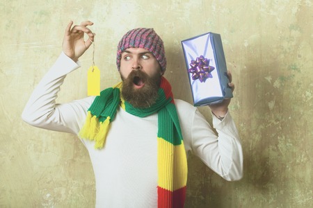 Hipster with present box in hat. Man with long beard in colorful scarf hold tag. Holiday celebration...