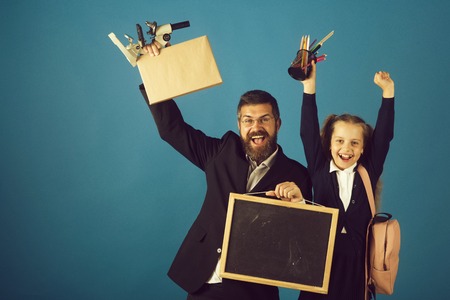 Kid and tutor hold school supplies and blackboard. Girl in uniform and bearded man in suit. Teacher and schoolgirl with cheerful faces on blue background. Classroom and alternative education conceptの写真素材