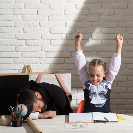 Schoolgirl with positive emotions and her sleeping tutor. Back to school and tiring homework concept. Girl and teacher in classroom on white brick background. Man and kid at desk with school suppliesの写真素材