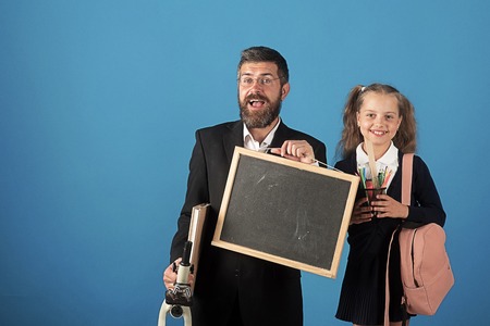 Kid and dad hold microscope, blackboard, book and stationery. Man and girl in school uniform. Home schooling and back to school concept. Father and schoolgirl with happy faces on blue backgroundの写真素材