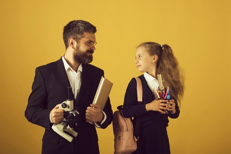 Girl with bag and bearded man. Classroom and alternative education concept. Teacher and schoolgirl with happy faces on yellow background. Kid and tutor hold book, pencils and microscopeの写真素材