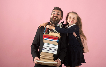 Girl in school uniform and bearded man in suit. Kid hugs her dad holding pile of books. Father and schoolgirl with happy faces on pink background. Home schooling and back to school conceptの写真素材
