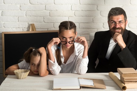 Kid and her tutors with tired, tricky and confident faces. Back to school concept. Teachers and schoolgirl on classroom background. Girl, lady and bearded man sit at school desk with booksの写真素材