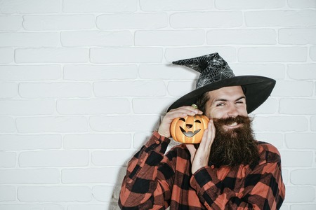 Halloween hipster with beard smiling in wizard hat. Jack o lantern. Holiday celebration concept. Trick or treat. Man with pumpkin on white brick wall, copy spaceの写真素材