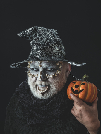 Wizard in witch hat on black background. Jack o lantern with spooky smile. Magic and halloween concept. Sorcerer with dragon skin and grey beard on face. Man with orange pumpkin.の写真素材