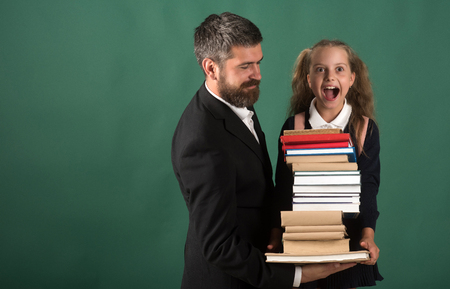 Girl in school uniform and bearded man. Kid and dad hold huge pile of books. Home schooling and back to school concept. Father and schoolgirl with excited faces on dark green background, copy spaceの写真素材