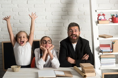 Girls and bearded man sit at desk with books. Kid, older sister and their tutor with happy faces. Home schooling and back to school concept. Teacher and schoolgirls on classroom backgroundの写真素材