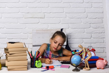Girl sits at desk with books stack, globe and colorful stationery. Schoolgirl with concentrated face writes in her copybook. Kid and school supplies on white brick background. Back to school conceptの写真素材