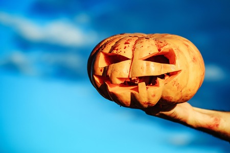 Halloween pumpkin with scary face, traditional autumn holiday symbol orange color with bloody male hand of zombie sunny outdoor on blue sky backgroundの写真素材