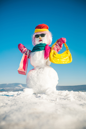 Christmas gift and xmas decoration. Snowman in pink wig, mittens, hat, glasses and scarf. Happy holiday and celebration. New year snowman from white snow with shopping bag. Winter fashion and party.の写真素材