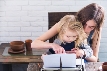 Sister teaching brother typing on vintage typewriter. Girl and baby at table on white brick wall....