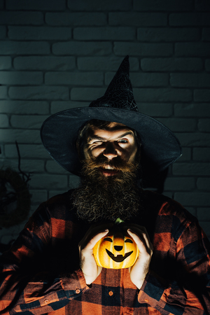Halloween hipster winking in witch hat and plaid shirt. Mystery and horror concept. Holiday celebration symbols on brick wall. Jack o lantern. Man with pumpkin in darkness.の写真素材