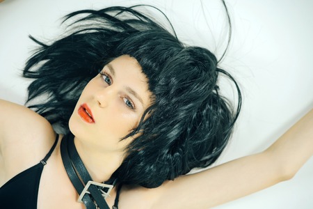 Model lying with leather belt on neck. Fashion accessories concept. Beauty and youth. Woman wearing black wig on grey background. Girl with red lips makeup on face.の写真素材