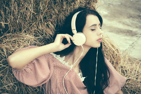 Girl listening music with closed eyes. Woman with headphones relaxing in hay. Leisure and entertainment. Summer vacation concept. Lifestyle and technology.の写真素材