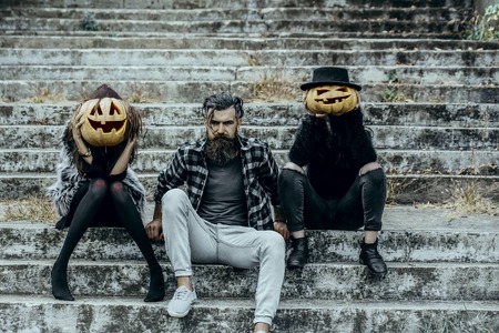 Halloween people or friends on stony stairs. Women with pumpkin heads. Bearded man sitting with girls. Halloween jack o lantern symbols. Holiday celebration concept.の写真素材