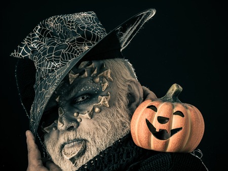 Man with orange pumpkin. Wizard in witch hat on black background. Jack o lantern with spooky smile. Magic and halloween concept. Sorcerer with dragon skin and grey beard on face.の写真素材