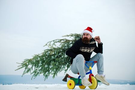 Cyclist in santa hat and sporty wear on winter day. Christmas and new year. Holiday celebration concept. Man riding tricycle on snowy landscape. Hipster carrying xmas tree on white snow.の写真素材
