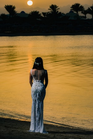 Girl in white dress on sunset sky, back view. Bride in wedding tiara on tropical landscape. Woman standing on sea beach. Honeymoon travel concept. Summer vacation and holidays.の写真素材