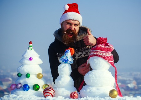 Santa man putting hat on snowman and showing ok gesture. Winter holidays celebration concept. Hipster in red santa claus cap. Christmas and new year decorations. Snow xmas tree and sculptures on blue sky.の写真素材