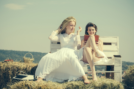 Brunette girl reading book. Blond woman putting straw wreath on head. Happy students sitting on bench on blue sky. Summer holidays and vacation. Education and knowledge concept.の写真素材