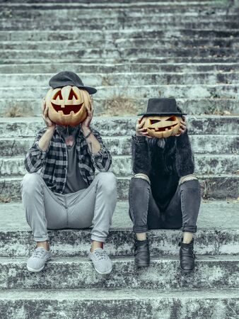 Halloween pumpkin woman in hat. Hipster couple sitting on grey stairs. Pumpkin man in cap. Halloween jack o lantern symbols. Holiday celebration concept.の写真素材