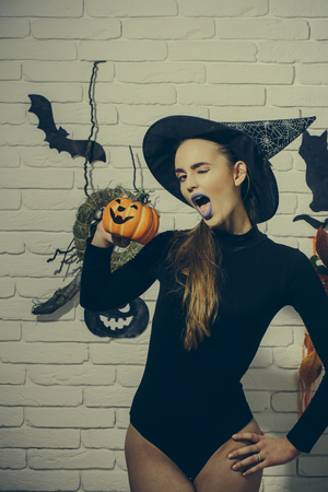 Halloween woman winking with blue lips and pumpkin. Holiday celebration symbols on white brick wall. Jack o lantern. Mystery and horror concept. Girl in witch hat and black bodysuit.の写真素材