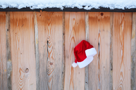 Santa claus hat on snowy fence. Red santa cap on wooden background. Winter palisade with white snow. Christmas and new year. Holidays celebration concept.の写真素材