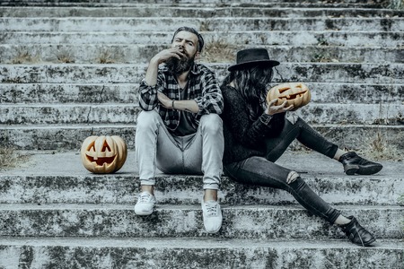 Halloween man and woman with pumpkins. Bearded hipster smoking cigarette. Halloween holiday celebration. Couple sitting on grey stairs. Bad habits and unhealthy lifestyle concept.の写真素材